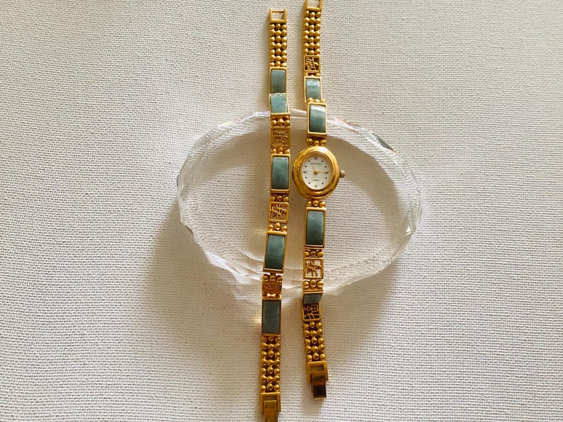 May include: A gold-toned wristwatch with a rectangular jade-colored stone bracelet. The watch face is white with black numerals and hands. The bracelet is made of gold-colored metal with jade-colored rectangular stones. The watch is displayed on a clear, oval-shaped glass.