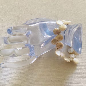 Può includere: Un espositore trasparente a forma di mano in plastica che tiene un braccialetto. Il braccialetto presenta più perline circolari piatte, bianche. L'espositore è su una superficie beige chiaro.