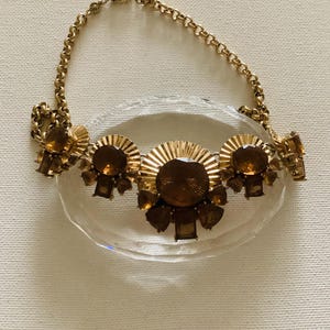 Könnte beinhalten: Eine goldfarbene Statement-Halskette mit bernsteinfarbenen Edelsteinen. Die Halskette hat eine Kette und ein zentrales Design mit einem Sonnenstrahlenmuster sowie rechteckigen und runden Steinen. Der Schmuck wird auf einer transparenten, ovalen Oberfläche präsentiert.