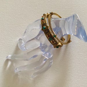 May include: A gold-toned metal bangle bracelet featuring a textured, rope-like edge and intricate filigree patterns. The bracelet is embellished with small, round, inlaid turquoise-colored stones and tiny red accents set along the outer surface.