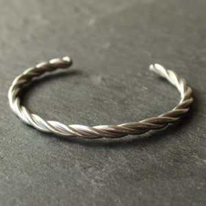 Könnte beinhalten: Ein silbernes, gedrehtes Armkettchen. Das Armband besteht aus zwei Silberdrähten, die miteinander verdreht sind, um ein seilartiges Design zu schaffen.