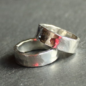 May include: Two silver rings with a hammered texture are stacked on top of each other. The rings have a flat band design and a shiny, reflective surface. The rings are likely made of sterling silver or a similar metal.