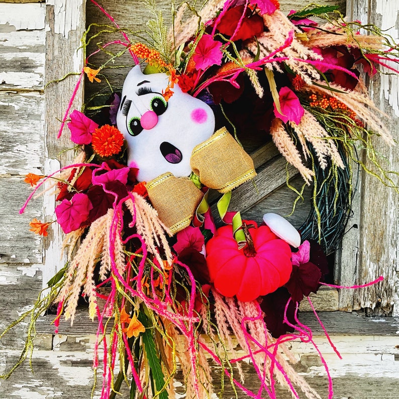 May include: A pink and white ghost-shaped door hanger with a burlap bow and pink flowers. The door hanger is decorated with pink and white feathers and a pink pumpkin.