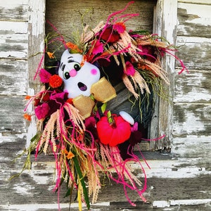 May include: A pink and white ghost-shaped door wreath with a pink and white floral arrangement. The wreath is decorated with pink, white, and green feathers and a pink and white bow.