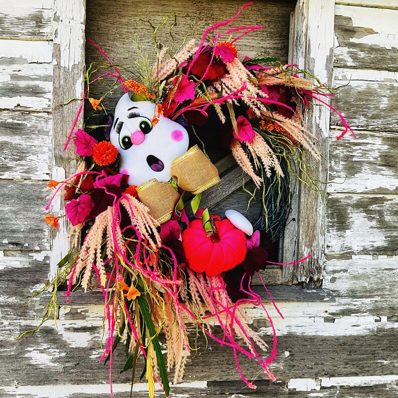 May include: A pink and white ghost-shaped door wreath with a pink and white floral arrangement. The wreath is decorated with pink, white, and green feathers and a pink and white bow.
