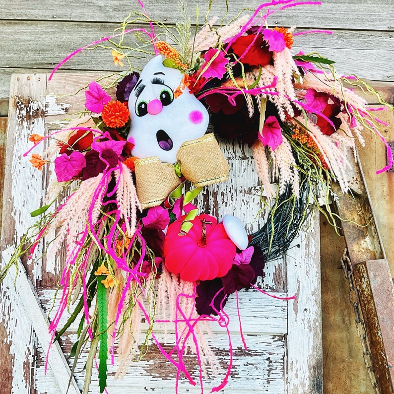 May include: A pink and white ghost face with a pink bow is the centerpiece of a fall wreath. The wreath is made of pink, red, and white flowers and greenery.