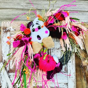 May include: A pink and white ghost with a bow tie is the centerpiece of a wreath made with pink, white, and green flowers. The wreath is decorated with pink and white feathers.