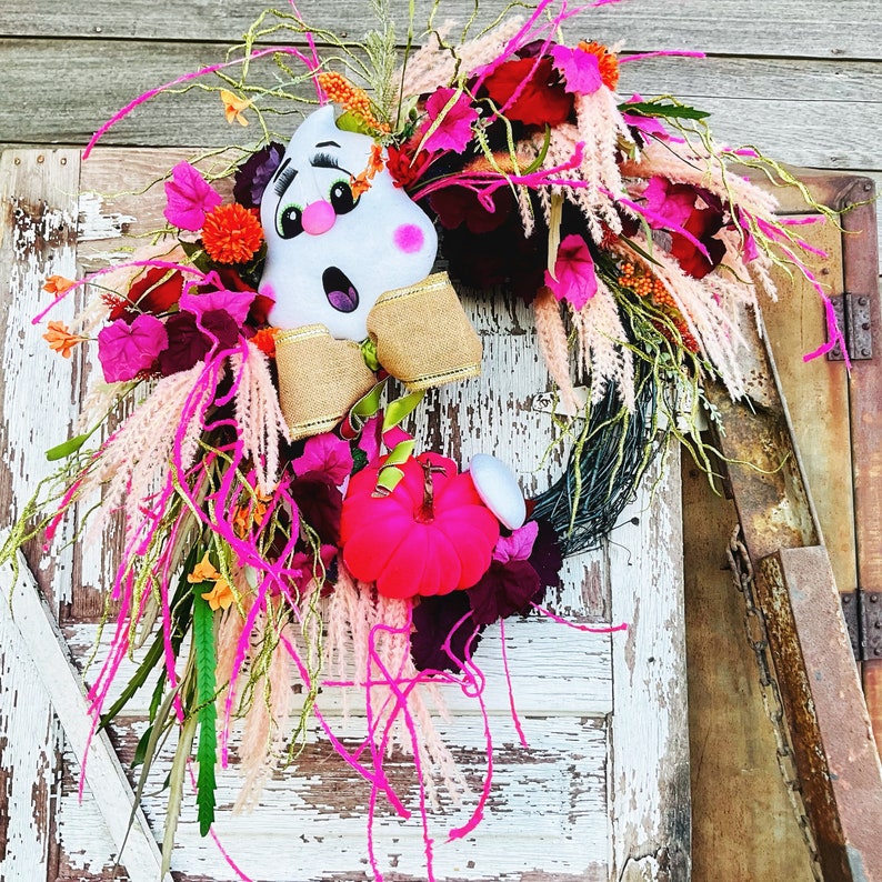 May include: A pink and white ghost with a black mouth and eyes is the centerpiece of a fall wreath. The wreath is made of pink, white, and green foliage and has a brown burlap bow.