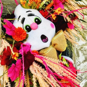 May include: A white ghost-shaped door hanger with pink cheeks and green eyes. The ghost is decorated with a gold bow and surrounded by pink, red, and orange flowers.