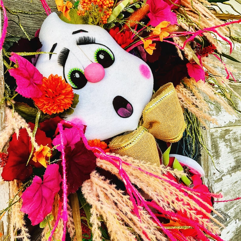 May include: A white ghost-shaped door hanger with pink cheeks and green eyes. The ghost is decorated with a gold bow and surrounded by pink, red, and orange flowers.