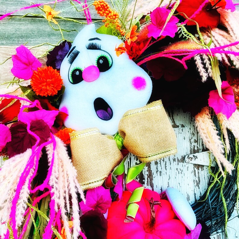 May include: A Halloween wreath featuring a white ghost with a pink nose and black eyes, wearing a brown and gold bow. The wreath is decorated with pink, red, and green flowers and greenery.