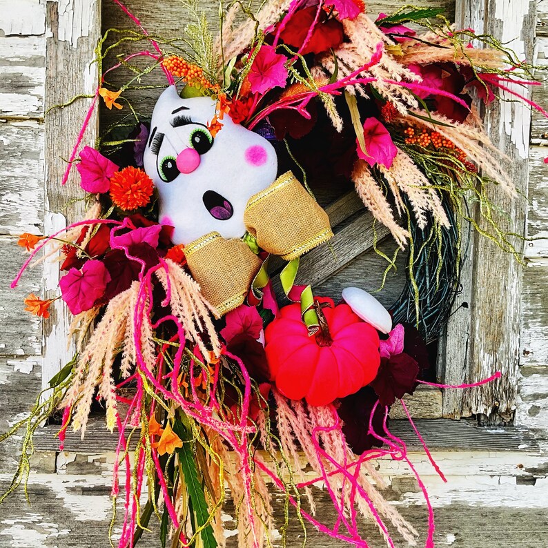 May include: A white ghost-shaped door hanger with a pink and gold bow, surrounded by pink, orange, and green fall foliage.