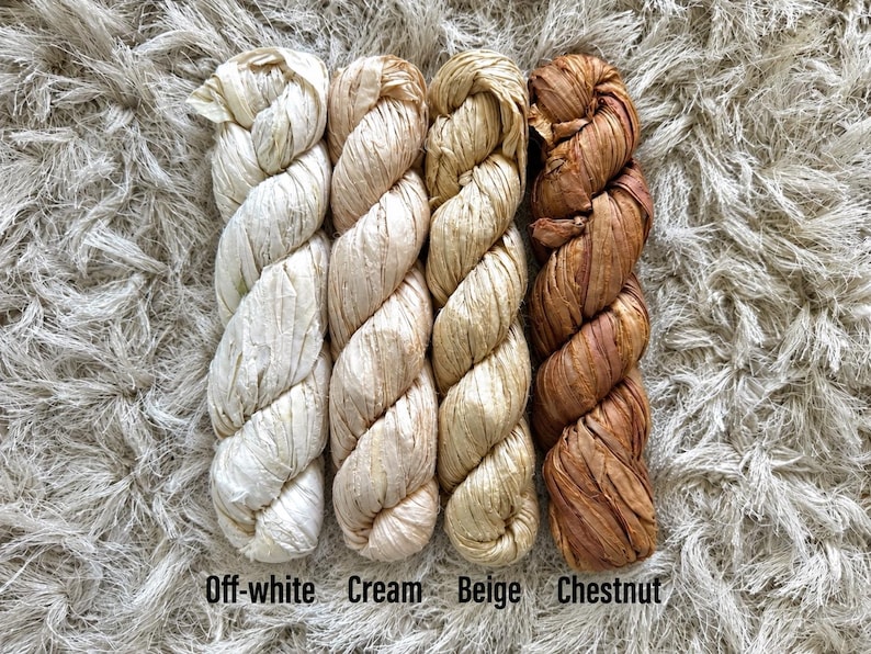 May include: Four twisted skeins of raffia ribbon arranged vertically. The colors from left to right are labeled as Off-white, Cream, Beige, and Chestnut. Each skein features a textured, fibrous appearance with a natural matte finish and consistent twist.