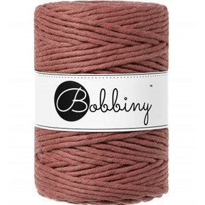 May include: A spool of dusty rose-colored macrame cord. The cord is tightly wound and wrapped with a white label featuring the word "Bobbiny" in a handwritten style font. The cord is suitable for various crafting projects.