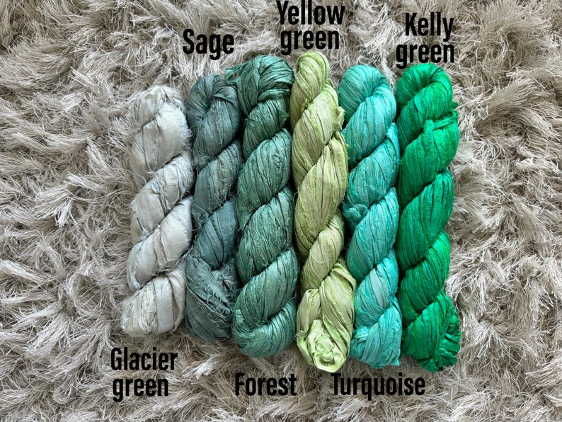 May include: Five skeins of textured ribbon yarn arranged side by side in a gradient of green shades. The colors are labeled as Glacier green, Sage, Forest, Yellow green, Turquoise, and Kelly green, showing a variety of light to dark tones.