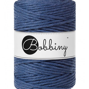 May include: A spool of blue macrame cord. The cord is tightly wound and has a textured appearance. A white label with the word "Bobbiny" in a script font is wrapped around the center of the spool.