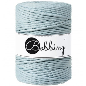 May include: A cylindrical spool of light blue macrame cord. The cord is tightly wound and wrapped with a white label featuring the word "Bobbiny" in a script font within a black circle. The cord appears soft and is suitable for various crafting projects.