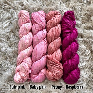 May include: Four skeins of textured ribbon yarn arranged side by side in a gradient of pink shades. The colors are labeled from left to right as Pale pink, Baby pink, Peony, and Raspberry. Each skein features a crinkled, matte fabric texture.
