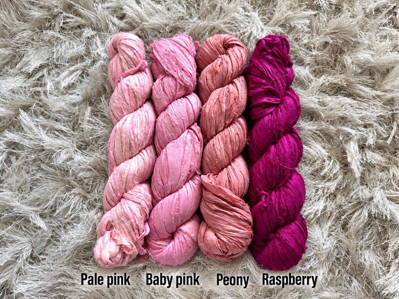 May include: Four skeins of textured ribbon yarn arranged side by side in a gradient of pink shades. The colors are labeled from left to right as Pale pink, Baby pink, Peony, and Raspberry. Each skein features a crinkled, matte fabric texture.
