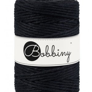 May include: A cylindrical spool of black macrame cord. A white label wraps around the center, displaying the brand name "Bobbiny" in a cursive font. The cord appears tightly wound and ready for crafting projects.