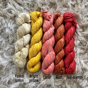 May include: Five skeins of ribbon yarn arranged side by side in a gradient of colors. The colors from left to right are labeled as pastel lemon, bright yellow, coral, rust, and red-orange. Each skein has a twisted, textured appearance.