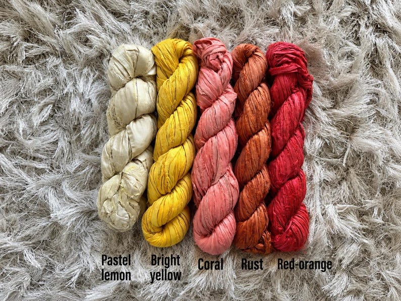 May include: Five skeins of ribbon yarn arranged side by side in a gradient of colors. The colors from left to right are labeled as pastel lemon, bright yellow, coral, rust, and red-orange. Each skein has a twisted, textured appearance.
