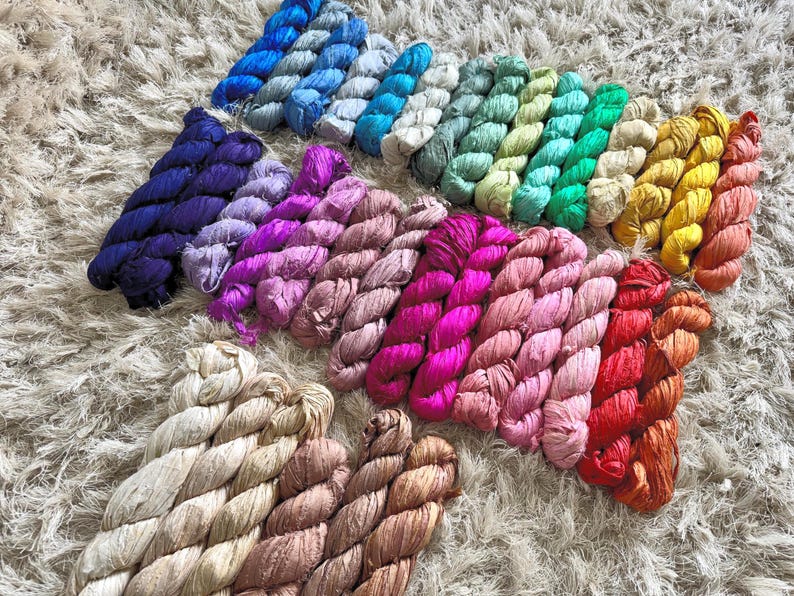May include: A collection of numerous small, twisted skeins of ribbon yarn arranged in a gradient pattern. The colors include shades of blue, green, yellow, orange, red, pink, purple, and neutral beige, all featuring a textured, crinkled appearance.