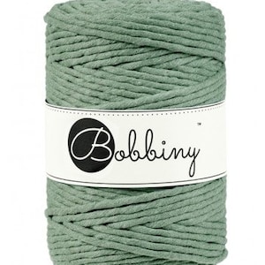 May include: A spool of sage green macrame cord. The cord is tightly wound and wrapped with a white label that reads "Bobbiny" in a script font. The cord is a solid color and appears to be made of cotton.