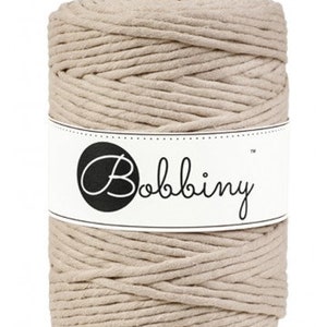 May include: A tightly wound spool of beige macrame cord. The cord is wrapped around itself, forming a cylindrical shape. A white label with the word "Bobbiny" in a script font is wrapped around the center of the spool. The label also features a black circle.