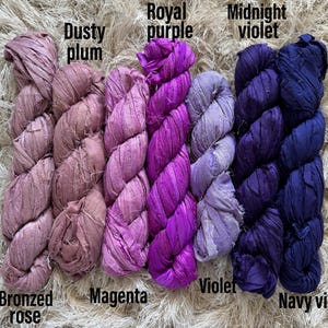 May include: Six skeins of crinkled silk ribbon yarn arranged in a row, labeled with the colors Bronzed rose, Dusty plum, Magenta, Royal purple, Violet, Midnight violet, and Navy violet. The ribbons have a textured, matte appearance.