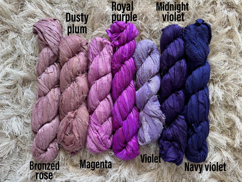 May include: Six skeins of crinkled silk ribbon yarn arranged in a row, labeled with the colors Bronzed rose, Dusty plum, Magenta, Royal purple, Violet, Midnight violet, and Navy violet. The ribbons have a textured, matte appearance.