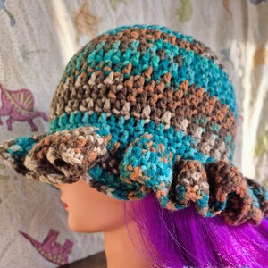 May include: A handmade crochet hat with a ruffled brim. The hat features a mix of teal, brown, beige, and orange yarn. The hat is designed with a rounded crown and a wide, wavy brim, offering a unique and stylish look.