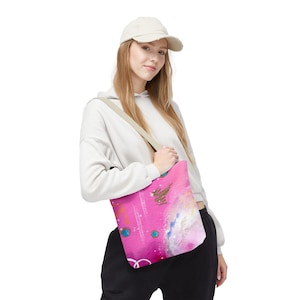 May include: A pink tote bag with an abstract design featuring gold, blue, and white accents. The bag has a beige shoulder strap. The bag is being carried by a person wearing a white hoodie and a cream-coloured baseball cap.