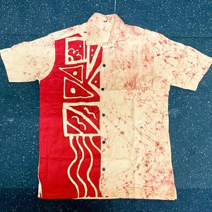 May include: Short-sleeved shirt with a distinctive pattern. The left panel is red with white geometric designs, the right is cream with red splatters. It has a collar and is fastened with black buttons.