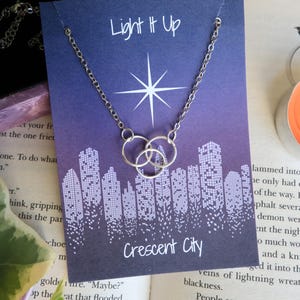 K&ouml;nnte beinhalten: Eine silberne Halskette mit zwei ineinander verschlungenen Kreisen h&auml;ngt an einer Karte mit violettem Hintergrund. Die Karte hat einen wei&szlig;en Stern und den Text "Light It Up" und "Crescent City".