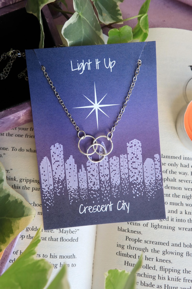 K&ouml;nnte beinhalten: Eine silberne Halskette mit zwei ineinander verschlungenen Kreisen h&auml;ngt an einer Karte mit violettem Hintergrund. Die Karte hat einen wei&szlig;en Stern und den Text "Light It Up" und "Crescent City".
