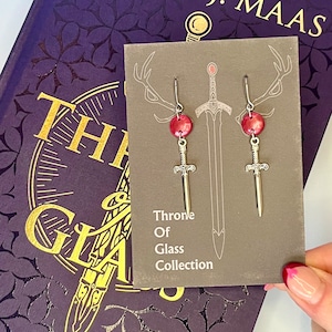 May include: A pair of silver sword earrings with red glass beads. The earrings are on a card that says "Throne of Glass Collection".