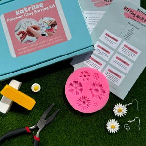Peut inclure: Un moule en silicone rose avec six formes de fleurs, une paire de boucles d'oreilles argentées avec des breloques de marguerite blanches, une barre d'argile jaune et blanche, une pince et une boîte bleue avec le texte "Katrilee Polymer Clay Earring Kit" sur le devant.