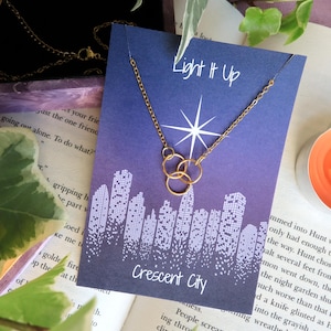 K&ouml;nnte beinhalten: Eine goldene Halskette mit drei miteinander verbundenen Kreisen h&auml;ngt an einer blauen Karte mit dem Text "Light It Up" und "Crescent City". Die Karte zeigt einen wei&szlig;en Stern und ein Stadtbild.