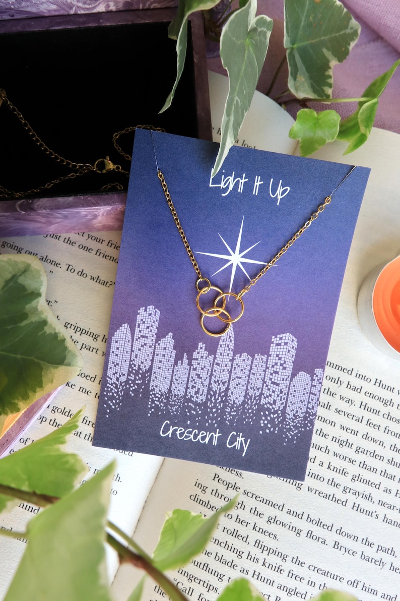K&ouml;nnte beinhalten: Eine goldene Halskette mit drei miteinander verbundenen Kreisen h&auml;ngt an einer blauen Karte mit dem Text "Light It Up" und "Crescent City". Die Karte zeigt einen wei&szlig;en Stern und ein Stadtbild.