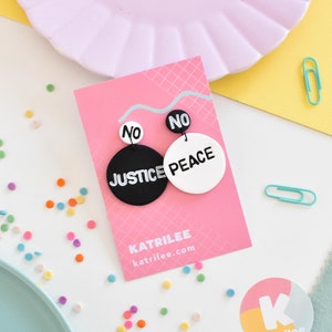 May include: A pair of black and white round earrings with the words "No Justice No Peace" on them. The earrings are displayed on a pink card with the text "Katrilee katrilee.com".