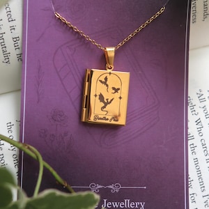 Puede incluir: Collar con colgante dorado con un medallón rectangular con siluetas de dragones y el texto "Breathe Fire". El medallón cuelga de una delicada cadena, exhibido en una tarjeta morada con la palabra "Jewellery".