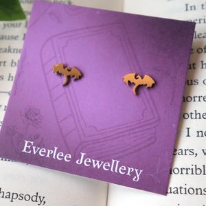 Può includere: Un paio di orecchini a forma di pipistrello color oro. Gli orecchini sono esposti su un cartoncino viola con la scritta "Everlee Jewellery". Il cartoncino è appoggiato su un libro aperto.