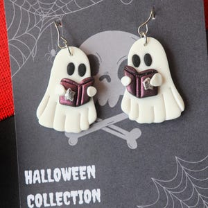 Op de afbeelding: Witte spookoorbellen met een paars boek met een ster. De oorbellen maken deel uit van de Halloween-collectie van Katrilee. De achtergrond is grijs met een schedel, kruisbeenderen en een spinnenweb.