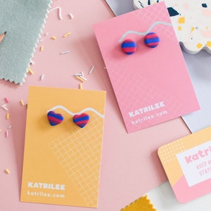 Puede incluir: Dos juegos de pendientes de arcilla polimérica de colores en tarjetas de presentación. Un juego presenta pendientes en forma de corazón con un patrón de rayas azul, rosa y rojo. El otro juego presenta pendientes redondos con un patrón de rayas similar. Ambas tarjetas presentan el texto "Katrilee katrilee.com".
