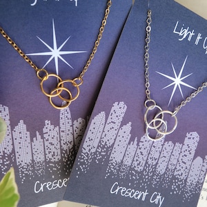 K&ouml;nnte beinhalten: Zwei Halsketten mit einem Stadtbild-Design und dem Text "Light It Up" und "Crescent City". Die Halsketten bestehen aus einer goldenen und silbernen Kette mit einem dreifachen Ring-Anh&auml;nger.