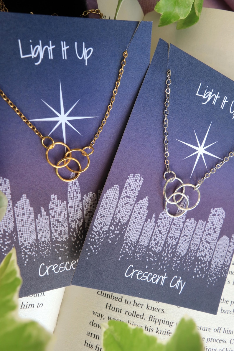K&ouml;nnte beinhalten: Zwei Halsketten mit einem Stadtbild-Design und dem Text "Light It Up" und "Crescent City". Die Halsketten bestehen aus einer goldenen und silbernen Kette mit einem dreifachen Ring-Anh&auml;nger.