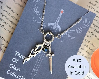 May include: A silver necklace with a sword, flame, and black gemstone pendant. The necklace is on a gray background with the text "Throne of Glass Collection" visible. Also Available in Gold.