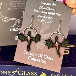 Puede incluir: Un par de pendientes de plata con alas de dragón negras y un corazón rojo. Los pendientes se muestran en una tarjeta con la cita "Somos los trece, desde ahora hasta que la oscuridad nos reclame." y el texto "Colección Throne of Glass".