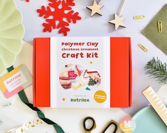Może przedstawiać: Czerwone pudełko z białą etykietą, na której widnieje napis "Polymer Clay Christmas Ornament Craft Kit" oraz "Created by Katrilee" i "Handmade in Britain". Pudełko otoczone jest materiałami do rękodzieła, w tym czerwoną śnieżynką, zieloną gałązką sosny i złotymi nożyczkami.